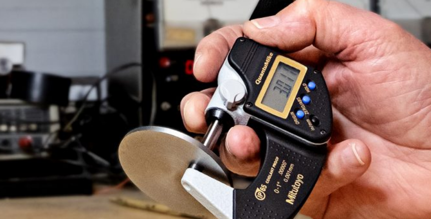 A person's hand holding a digital micrometer measuring the thickness of a metal disc in a workshop.