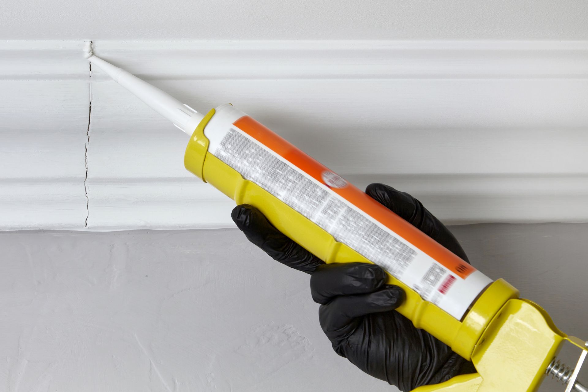 Hand with black glove, using a caulk gun to seal a crack in white molding.
